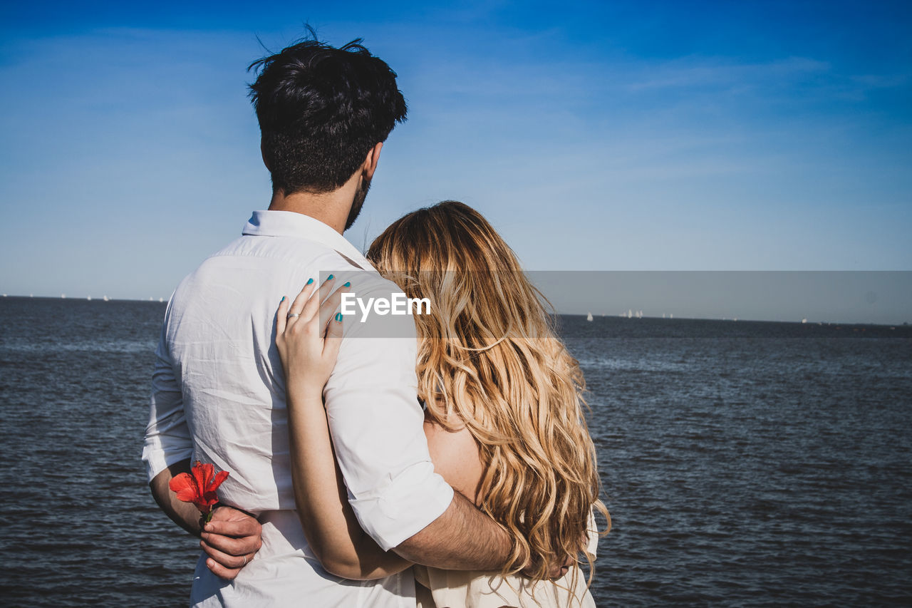 Rear view of couple with arm around standing by sea against sky