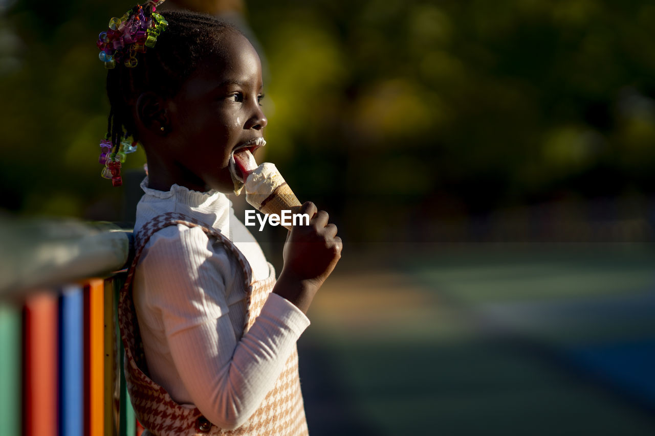 Side view of cheerful african american girl standing near colorful fence and licking sweet ice cream on street against blurred background