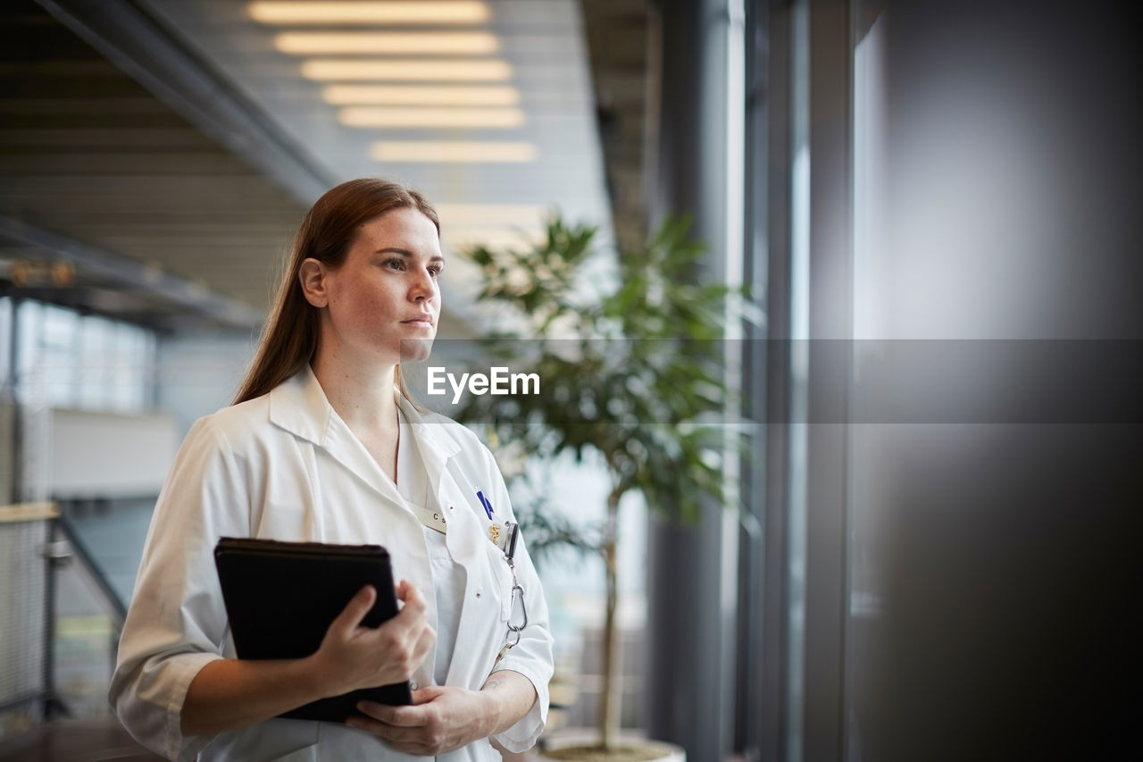 Thoughtful young female doctor with digital tablet looking through window at corridor in hospital