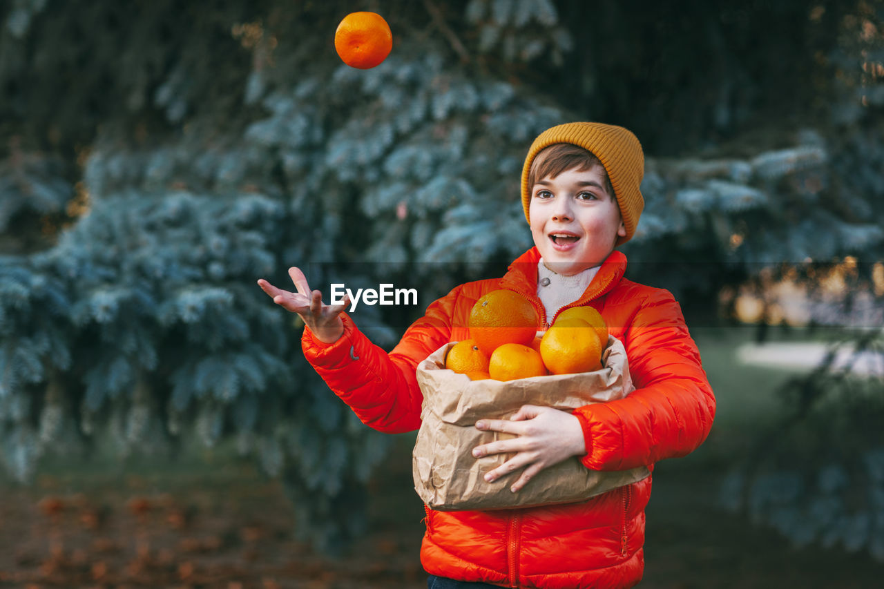 A boy in a bright orange jacket and yellow hat holds a large bag of oranges in his hands