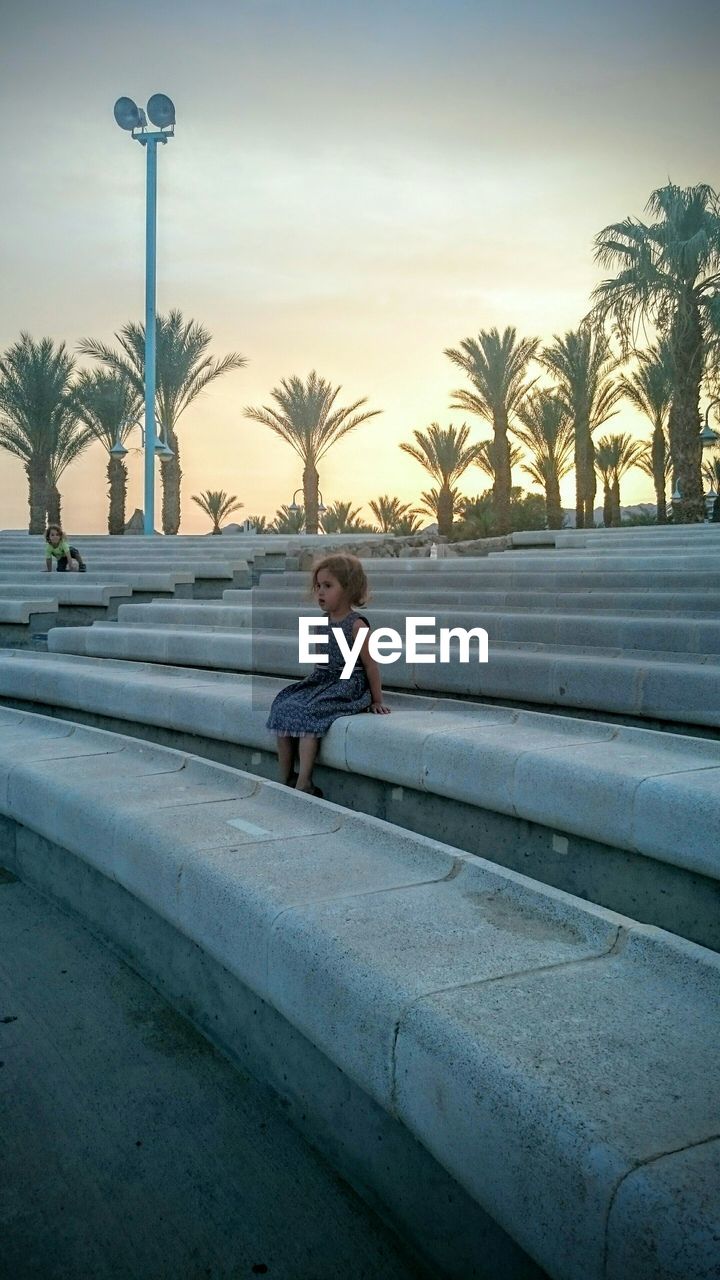 Children sitting on stone seats against sky at sunset