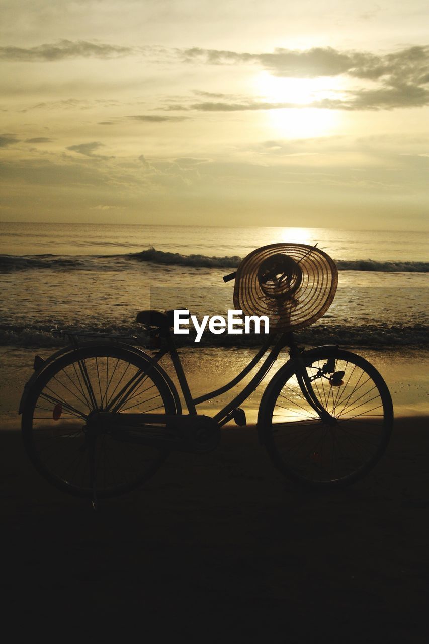 Bicycle with hat at beach against sky during sunset