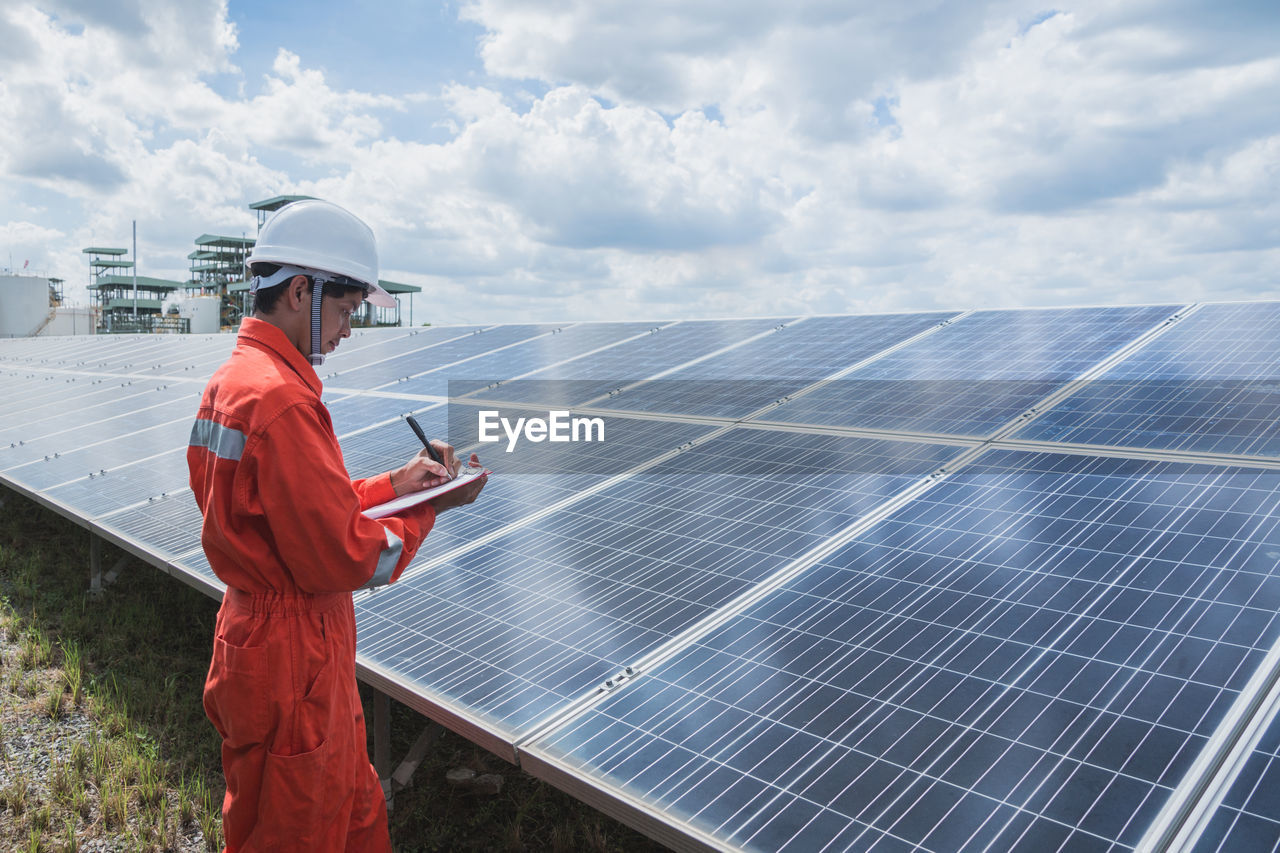 Engineer standing amidst solar panel against cloudy sky