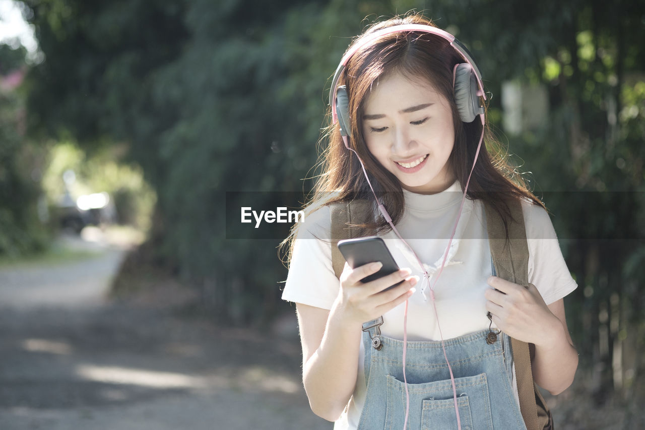 Smiling young woman listening music through headphones while standing against trees