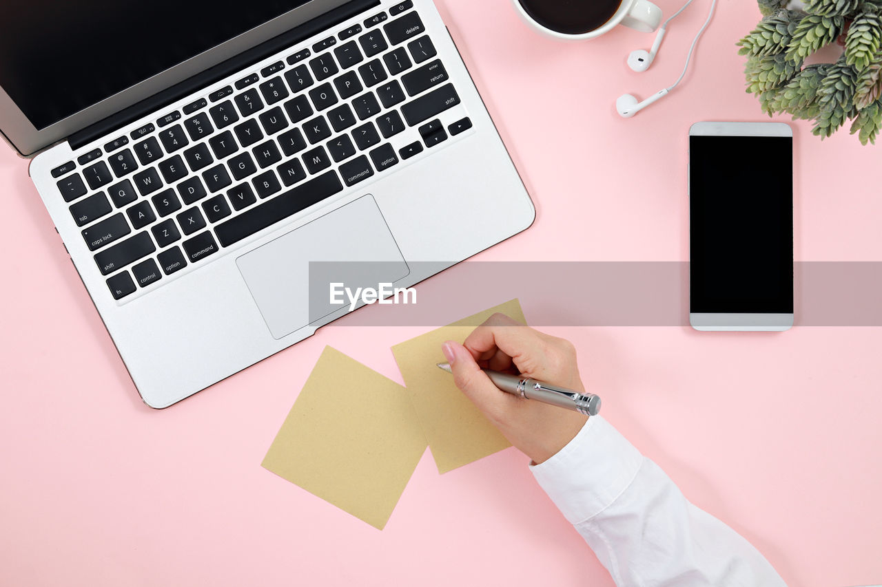 Directly above shot of businesswoman writing on adhesive note by laptop on pink background