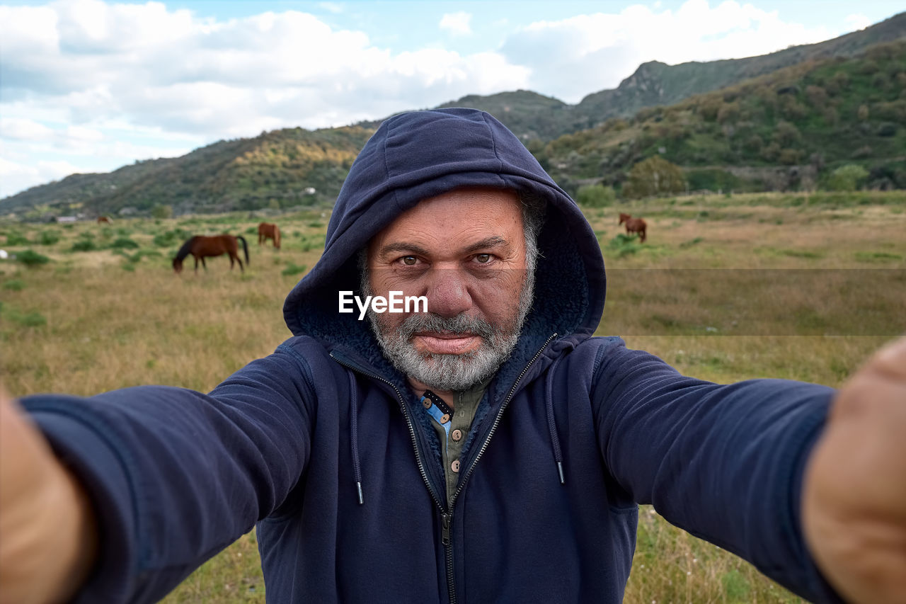 Happy, serious mature bearded man making a selfie on meadow with grazing horses. 