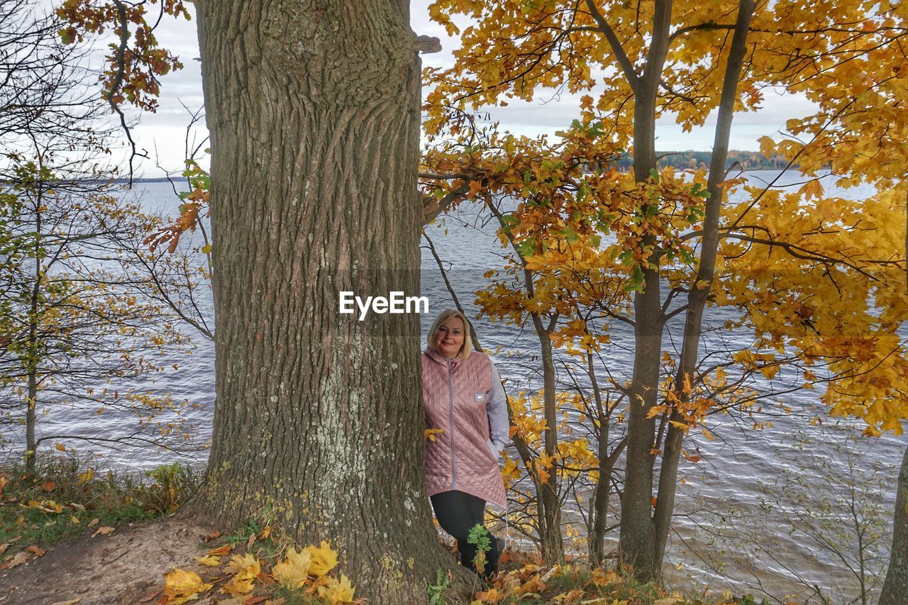Woman standing by tree trunk against lake 
