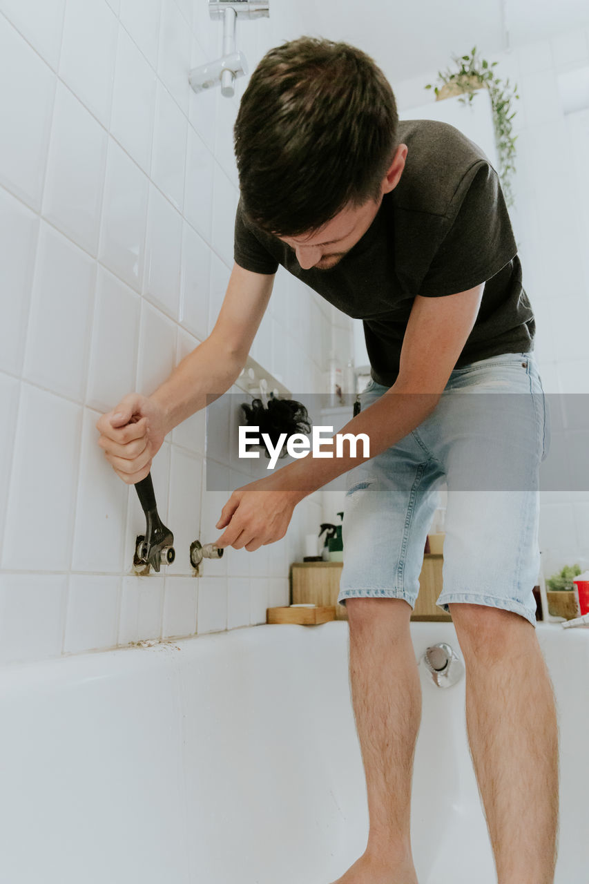A young man tightens the nut of a faucet in the wall with an adjustable wrench.