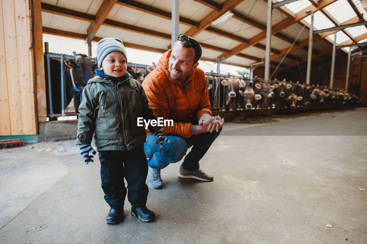Father and son smiling in barn with cows behind during winter