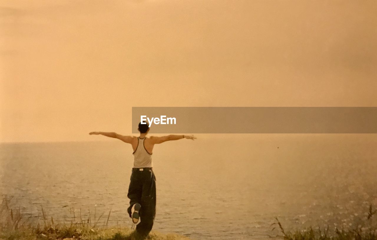 REAR VIEW OF MAN WITH ARMS OUTSTRETCHED STANDING ON SEA SHORE