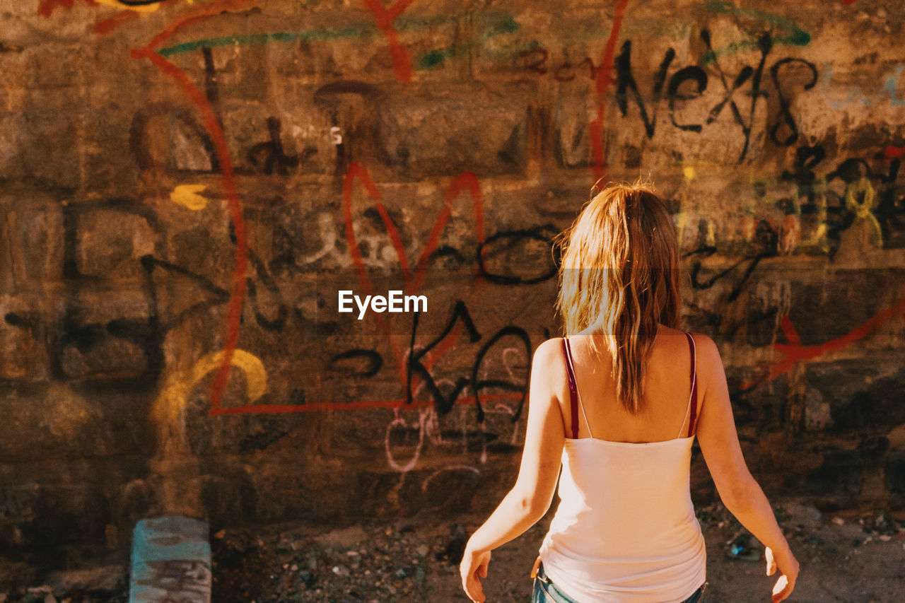 Rear view of woman standing against graffiti wall on sunny day