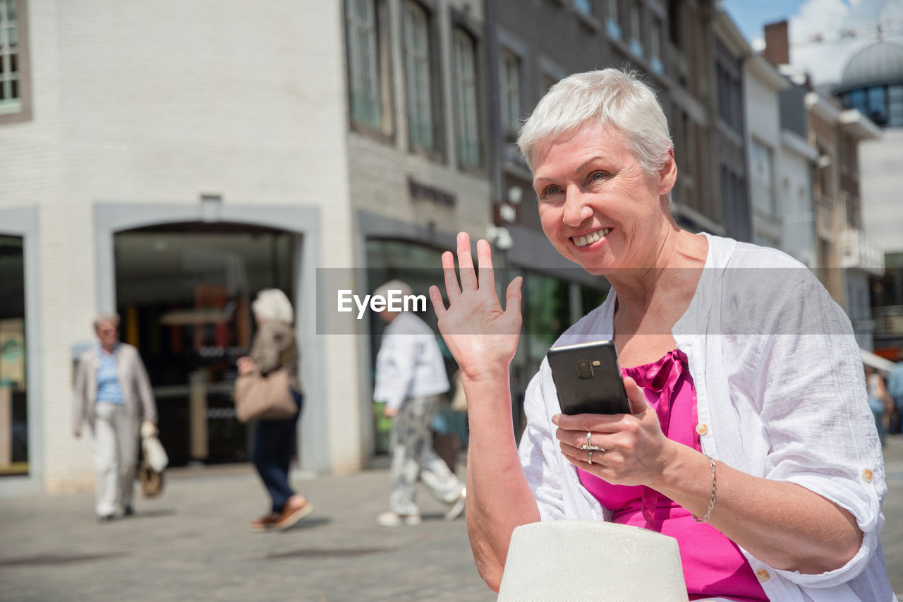 Sixty year old caucasian woman in stylish summer clothes waves to a passerby, holds smartphone