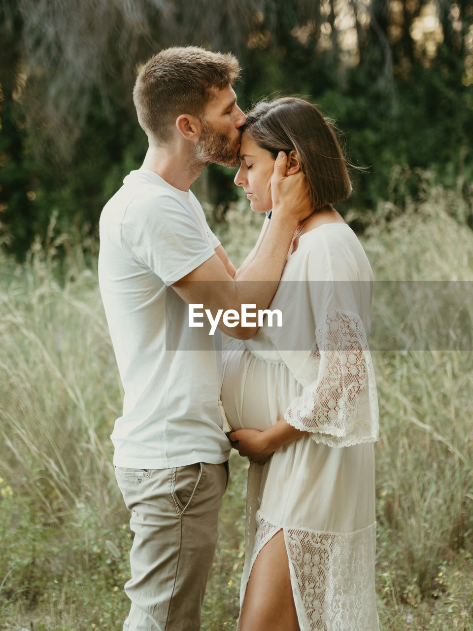 Side view of tender male kissing pregnant female in forehead while standing in field in nature