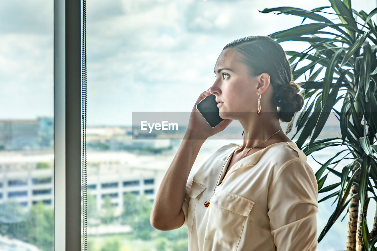 Woman talking on mobile phone against glass window in office