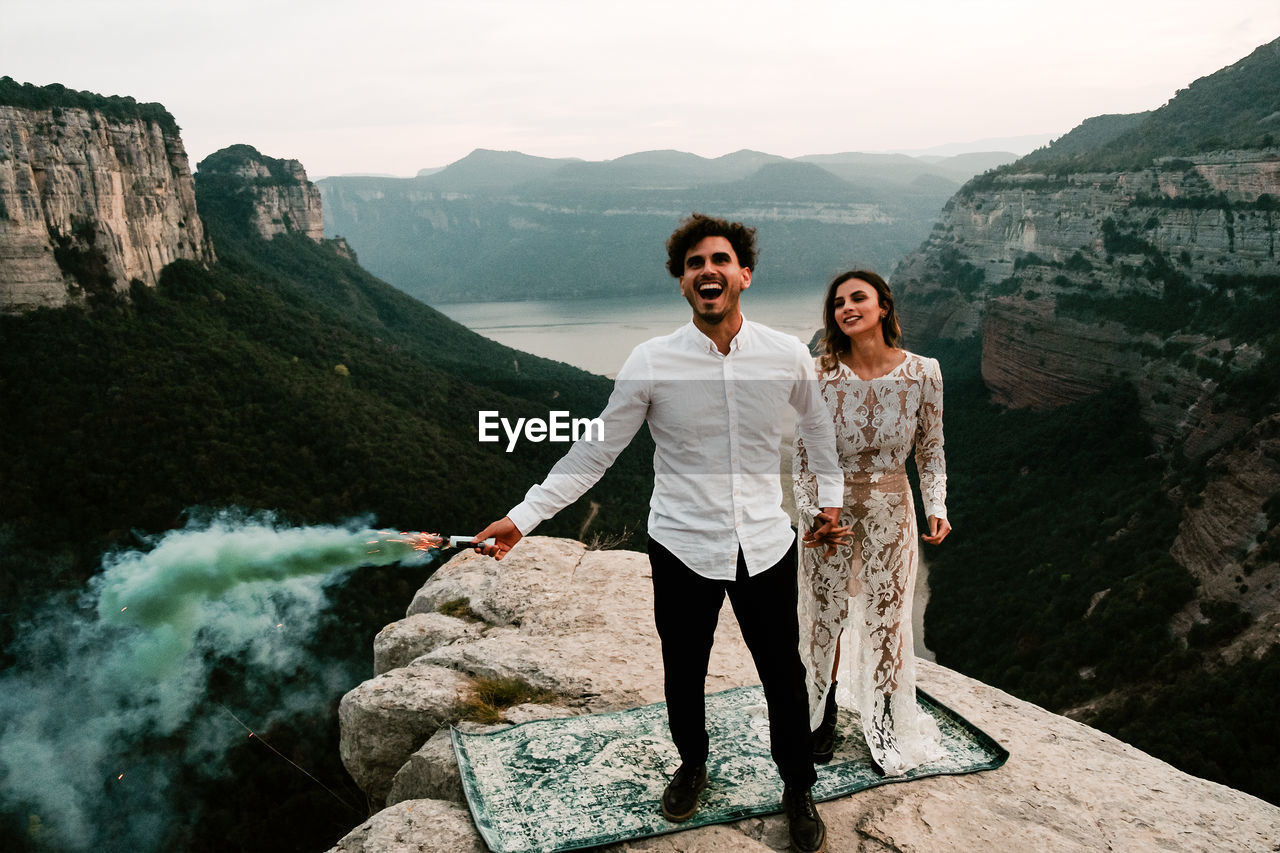 Full body of happy young newly married couple in elegant outfits holding colorful smoke bombs holding hands while celebrating marriage on top of rock in morro de labella in spain