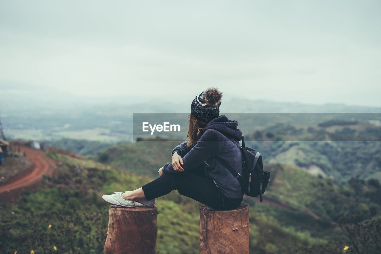 Side view of woman with backpack looking at landscape while sitting on wooden posts against sky