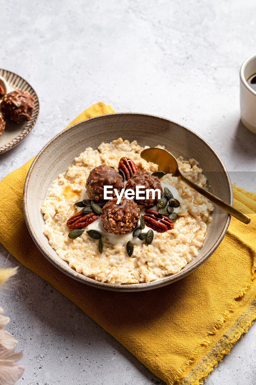 High angle of bowl with tasty milk oatmeal served with crunchy chocolate balls pumpkin seeds and pecan nut on napkin for healthy breakfast