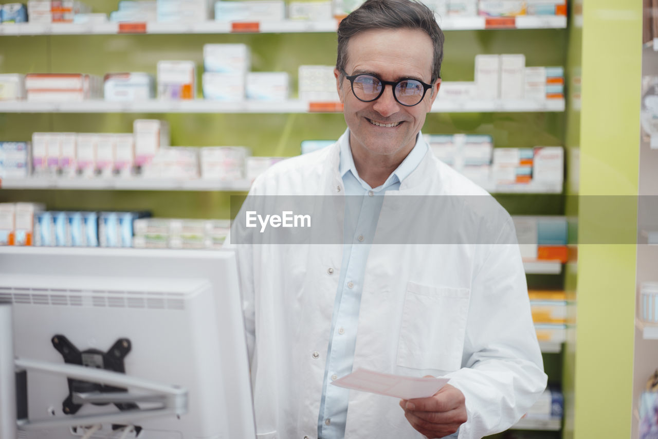 Happy pharmacist holding prescription in front of desktop pc at store