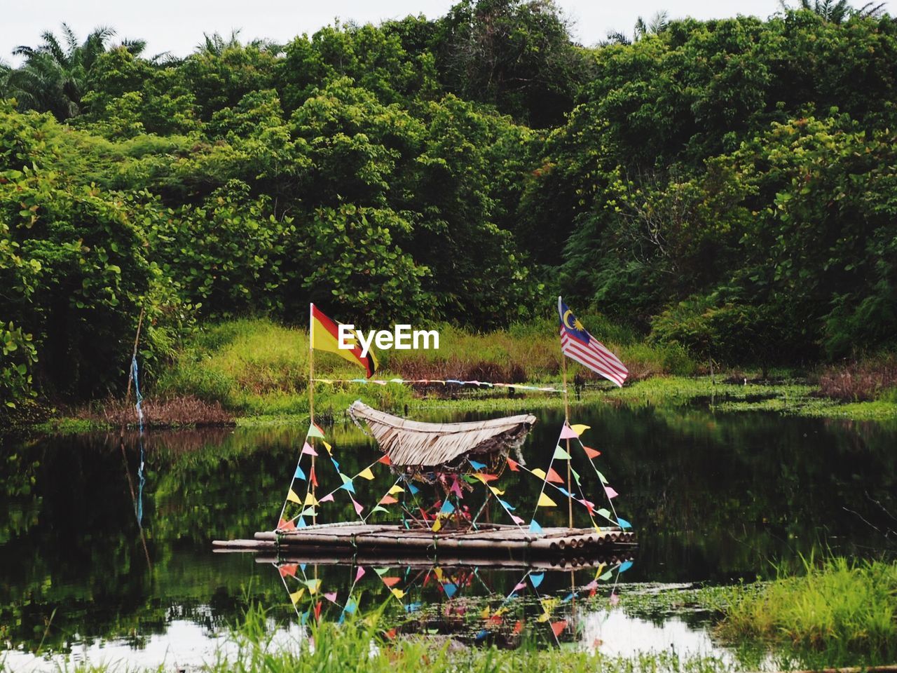 Flags and decoration on wooden raft moored at lake against trees