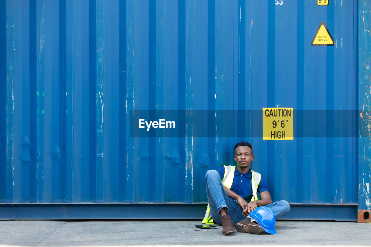 PORTRAIT OF SMILING YOUNG MAN SITTING AGAINST BLUE WALL