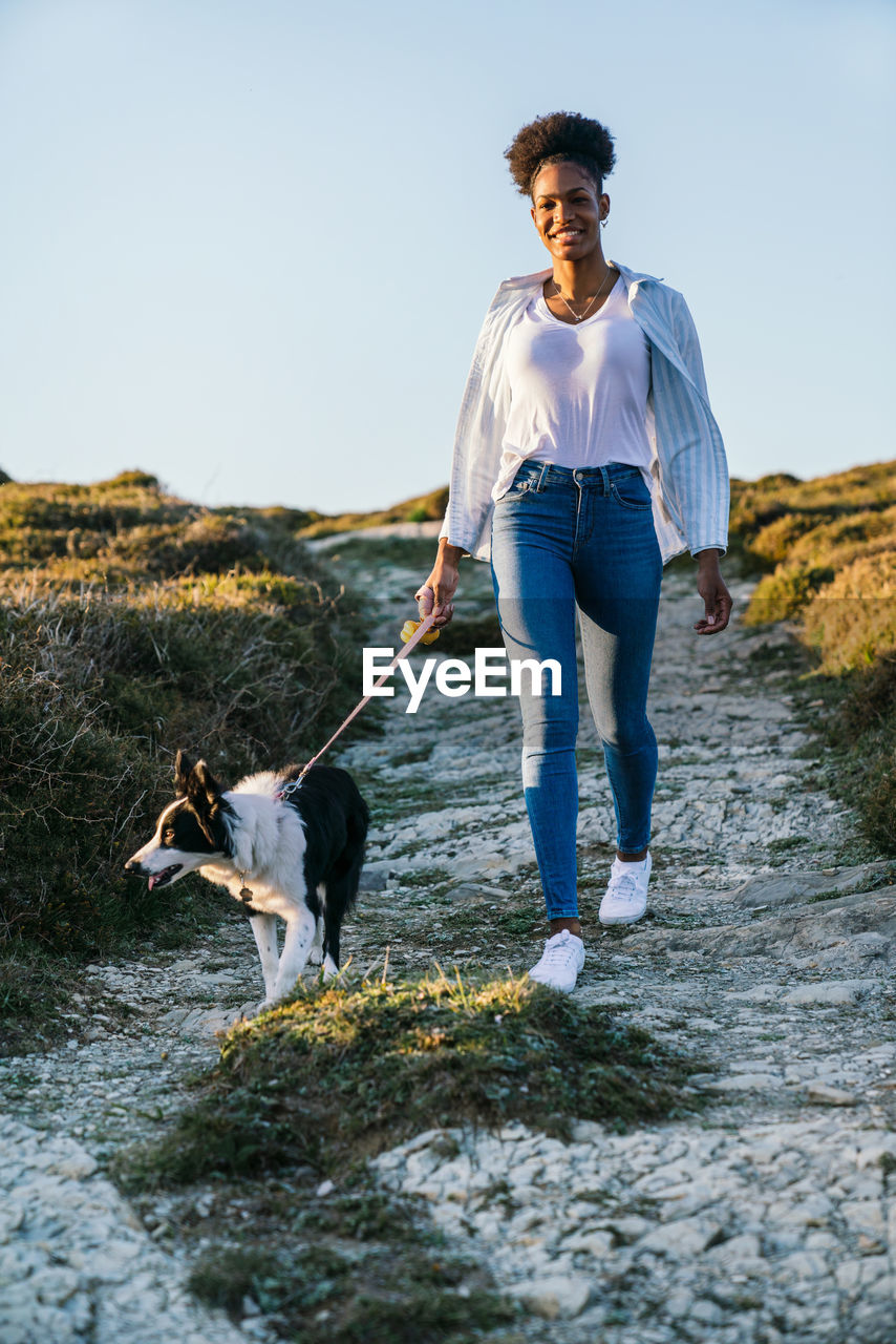 Full body of happy ethnic woman with border collie dog walking together on trail among grassy hills in sunny spring evening