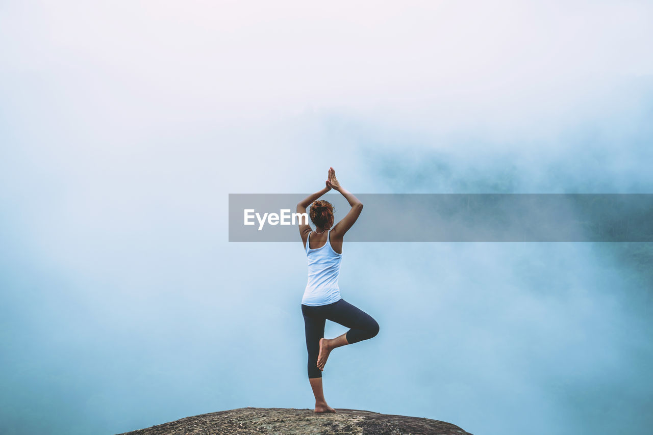 Full length rear view of woman with arms raised doing yoga o rock against sky