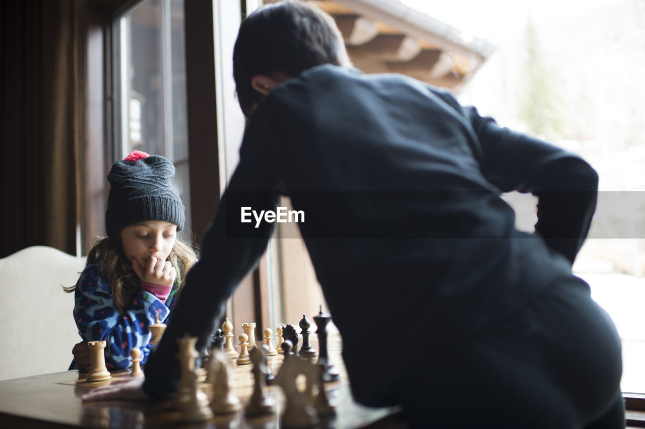 Siblings playing chess while standing by window at home