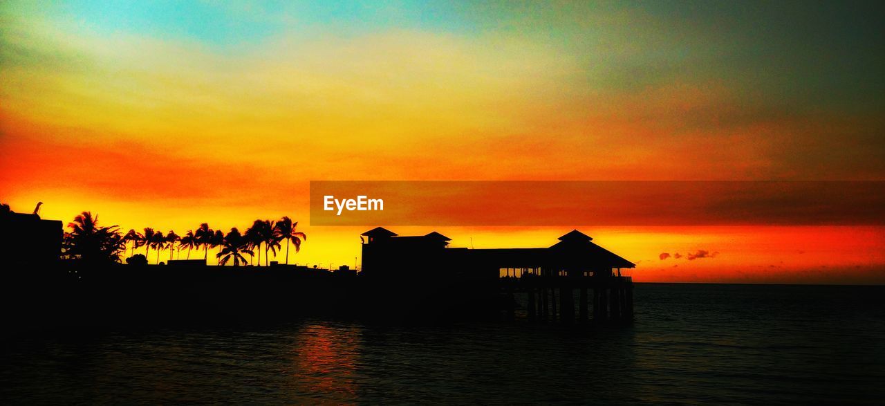 Silhouette stilt house by lake against sky during sunset