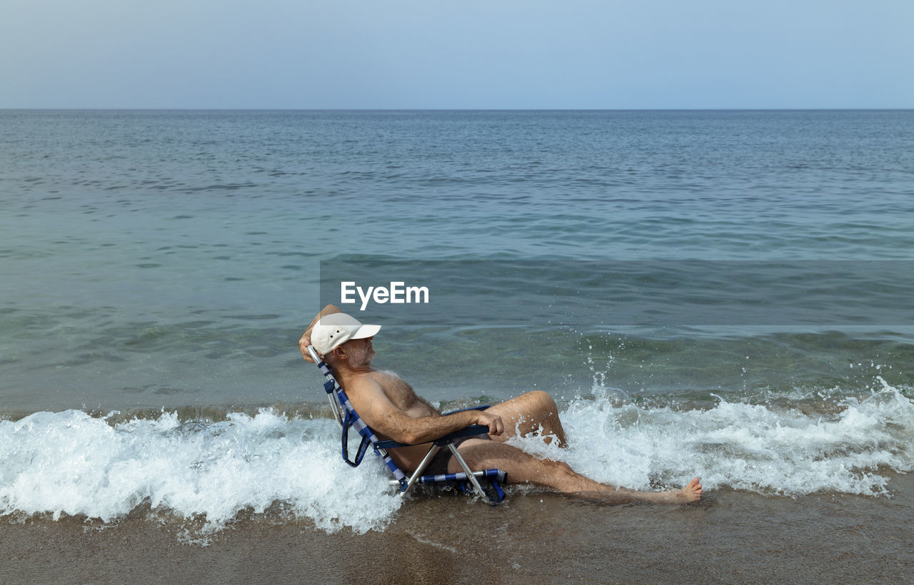 Side view of shirtless adult man in swimwear siting on chair on beach