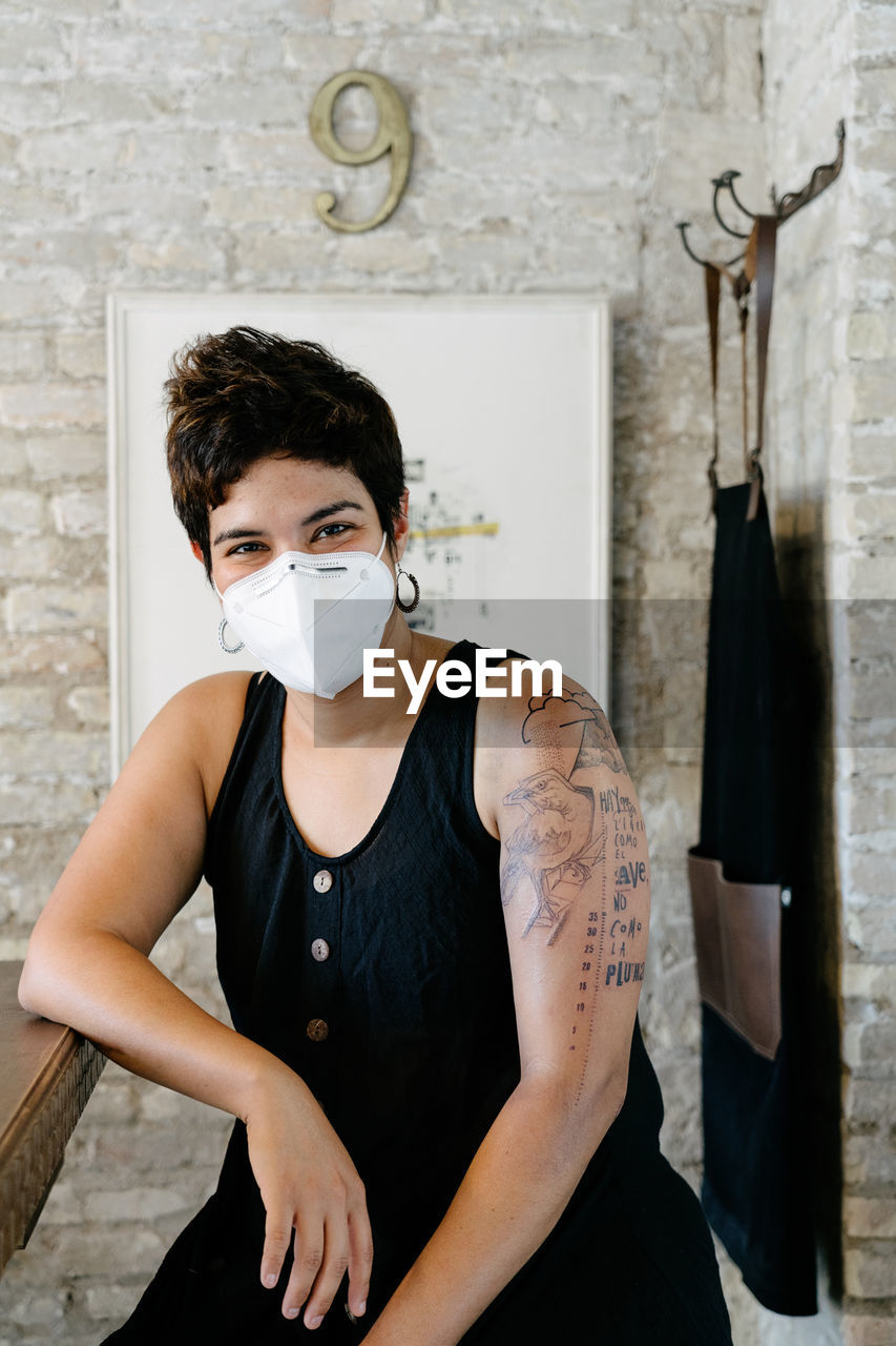 Delighted female in protective mask sitting at table in modern salon after procedure and enjoying new tattoo on arm while looking at camera