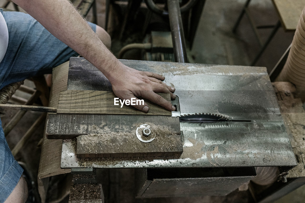 High angle of male carpenter sitting at workbench and cutting piece of wooden plank with sharp circular saw while working in shabby workshop