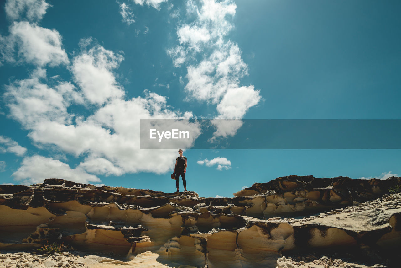 Low angle view of mature woman standing on rock against blue sky during sunny day