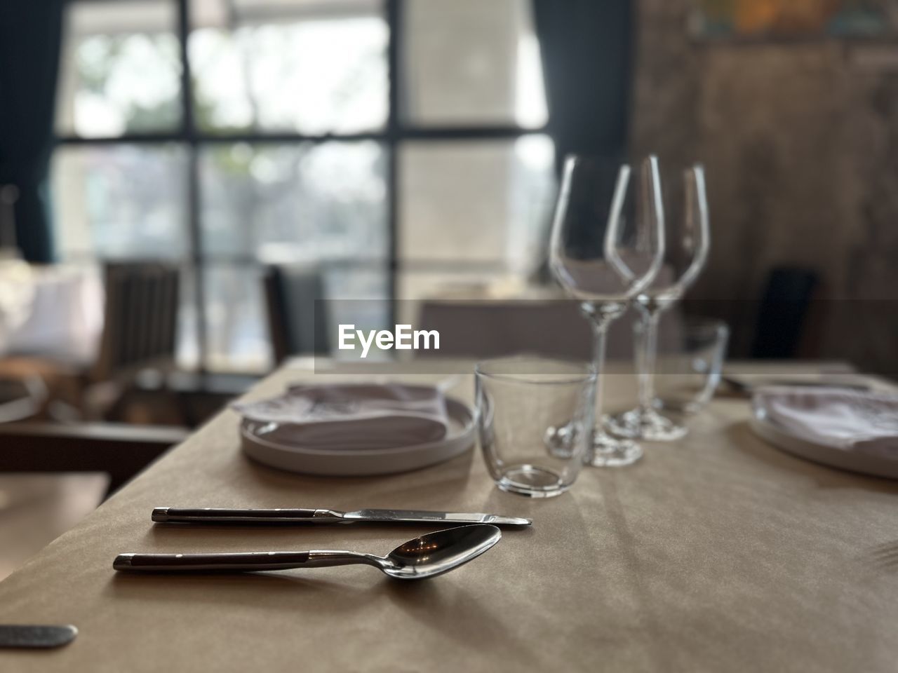 table, glass, setting, household equipment, eating utensil, place setting, kitchen utensil, fork, indoors, drinking glass, wine glass, furniture, dining table, food and drink, table knife, napkin, knife, business, restaurant, plate, no people, silverware, crockery, room, spoon, absence, empty, elegance, focus on foreground, drink, dining, empty plate, arrangement, meal, tablecloth, luxury, dining room, wealth, day, window, seat, domestic room, food, food and drink industry, wood, chair, selective focus, close-up, anticipation, still life, refreshment