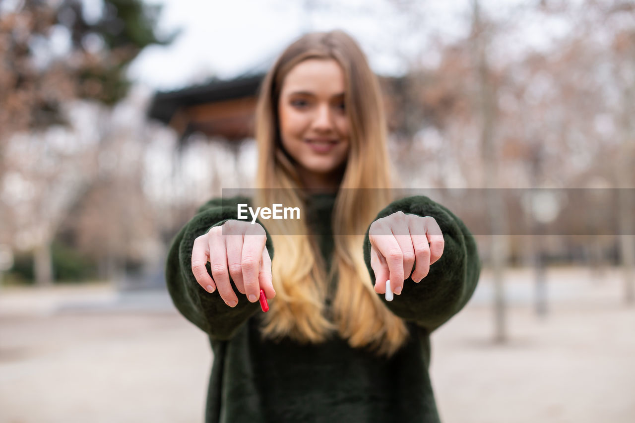 PORTRAIT OF SMILING YOUNG WOMAN STANDING AGAINST BLURRED BACKGROUND