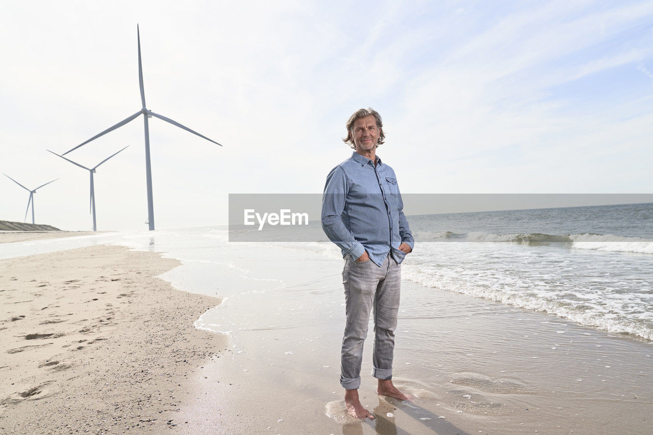 Smiling senior businessman standing in front of wind turbines at beach