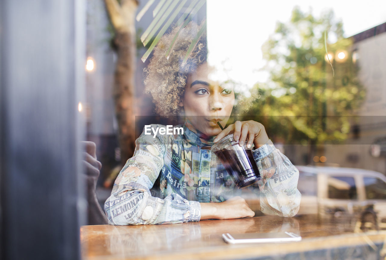 Woman drinking ice tea while sitting at cafe seen through glass window