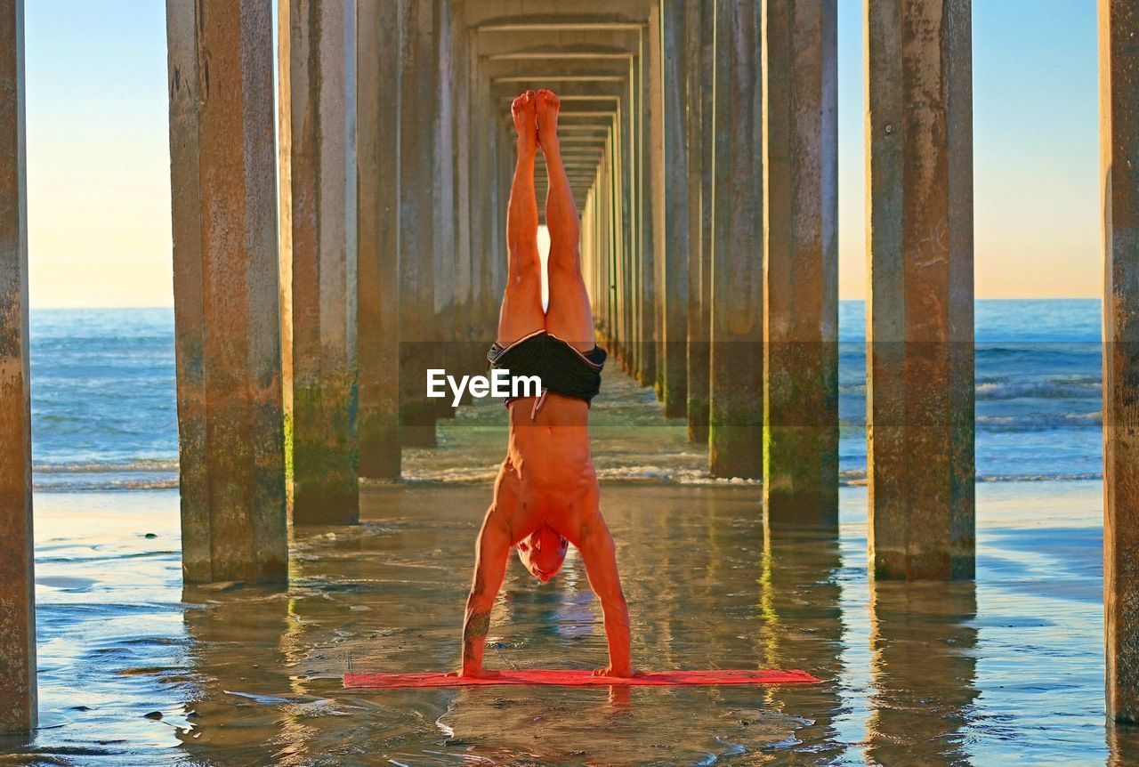 Shirtless man practicing yoga amidst architectural columns of pier at beach