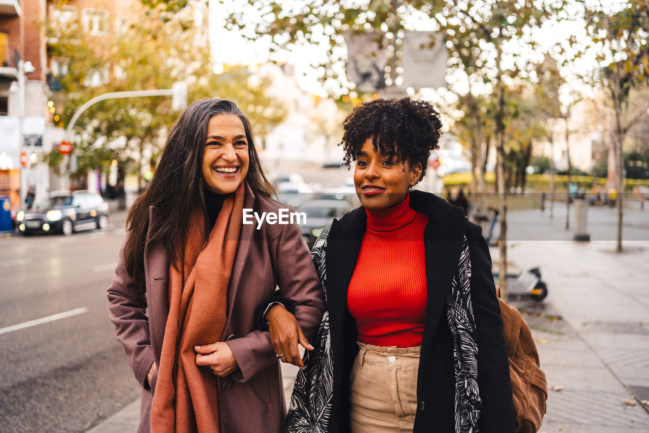 Cheerful lady holding arms black female friend in warm clothes while walking together on street pavement looking away