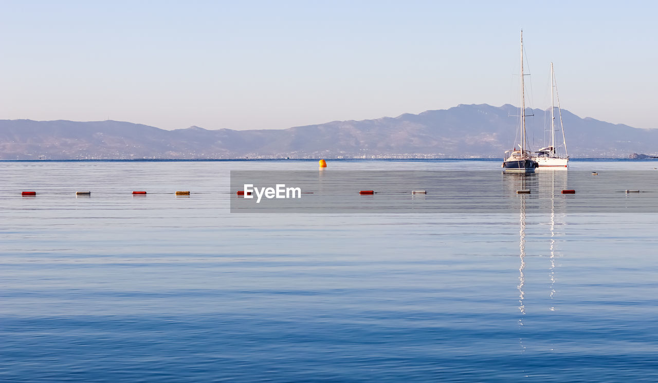 water, nautical vessel, transportation, sea, mode of transportation, sky, sailboat, boat, nature, vehicle, beauty in nature, mountain, buoy, boating, scenics - nature, clear sky, blue, tranquility, travel, day, bay, ship, no people, tranquil scene, sailing, pole, outdoors, travel destinations, watercraft, reflection, idyllic, waterfront, copy space, environment, sunny, mast, holiday, shore, non-urban scene, vacation, land, landscape, floating, sunlight, trip