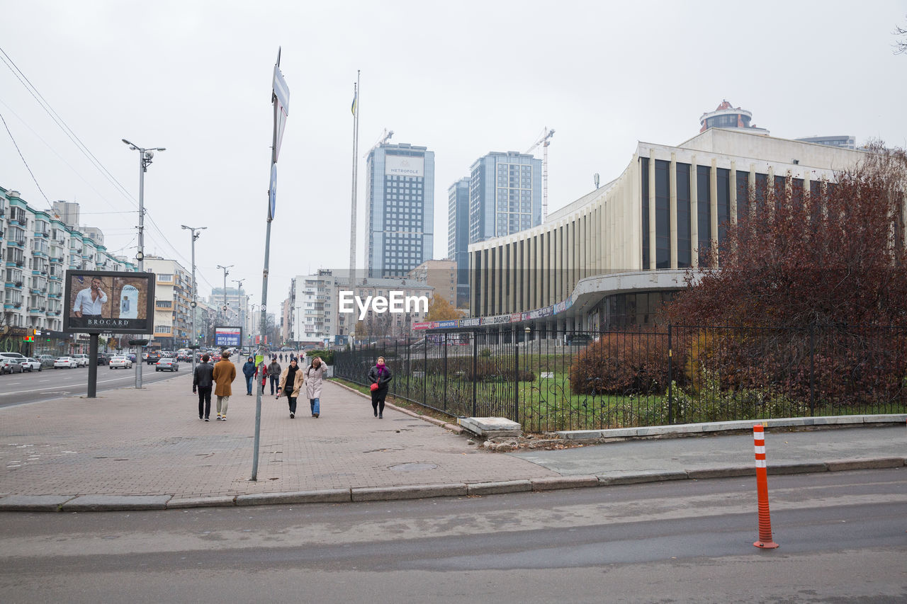 PEOPLE WALKING ON ROAD BY BUILDINGS AGAINST SKY