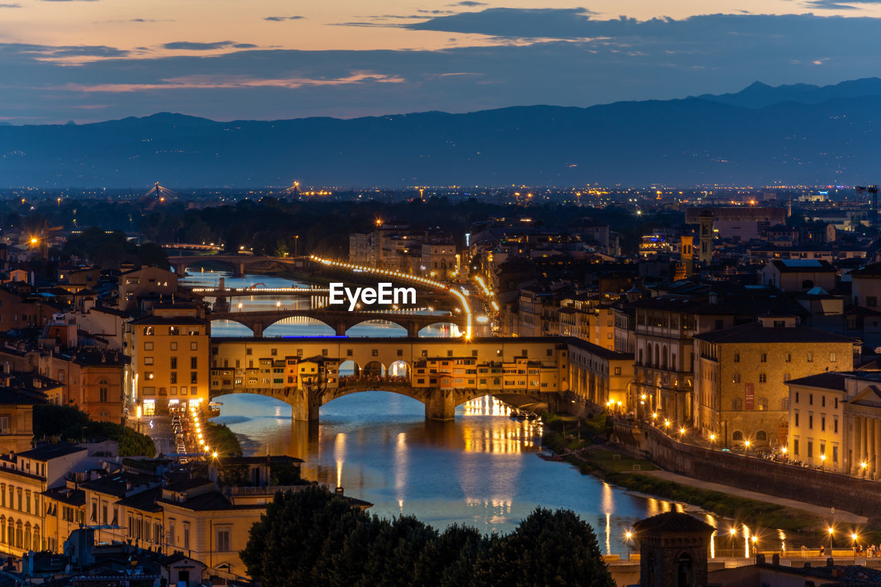 ILLUMINATED BRIDGE OVER RIVER AGAINST BUILDINGS IN CITY