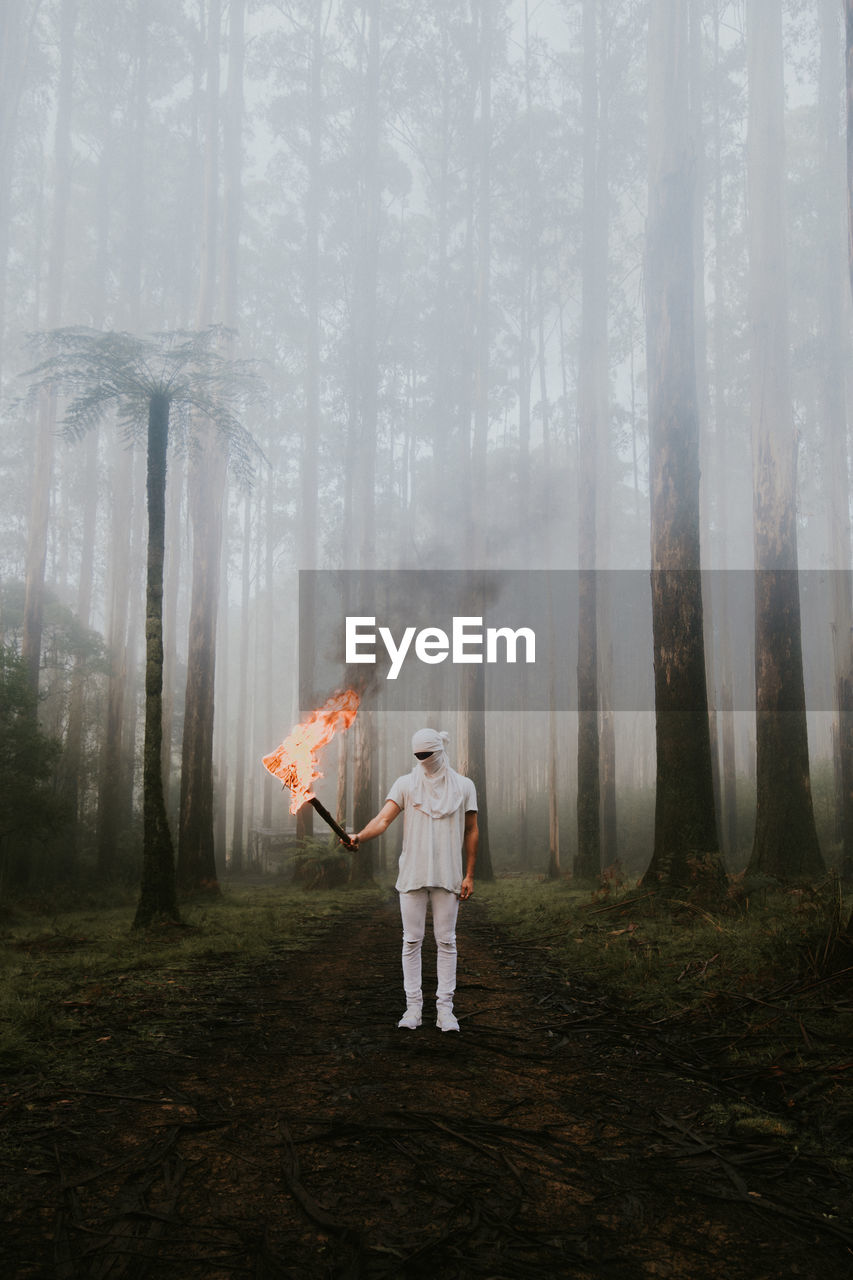 Full length of young man with fire standing in forest during foggy weather