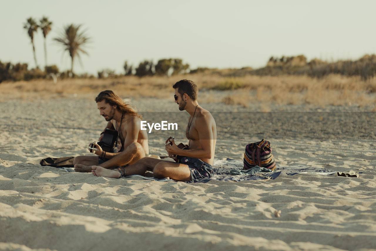 Relaxed talented man playing acoustic guitar for male friend while sitting together on sandy beach at sunset