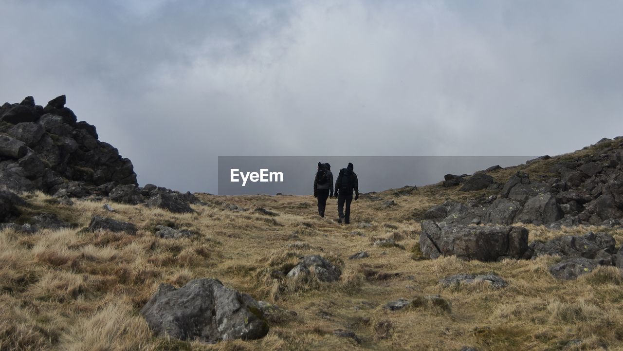 REAR VIEW OF MEN STANDING ON ROCK AGAINST SKY