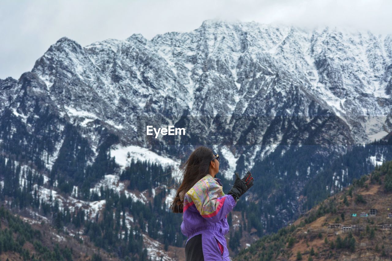 mountain, mountain range, leisure activity, beauty in nature, snow, one person, scenics - nature, winter, women, activity, nature, cold temperature, adult, ridge, environment, vacation, landscape, lifestyles, trip, adventure, hiking, day, holiday, snowcapped mountain, travel, standing, sky, outdoors, female, land, rear view, travel destinations, tranquil scene, backpack, mountaineering, tranquility, summit, backpacking, non-urban scene, clothing, young adult, tree, plant, sports, tourist, warm clothing, tourism, hairstyle, three quarter length, forest, walking, exploration, waist up, cloud, casual clothing