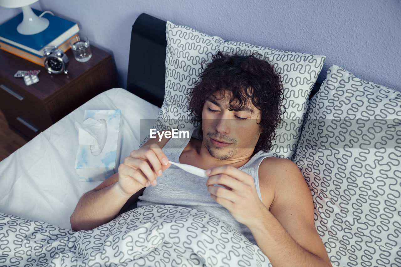 High angle view of young man holding thermometer on bed at home