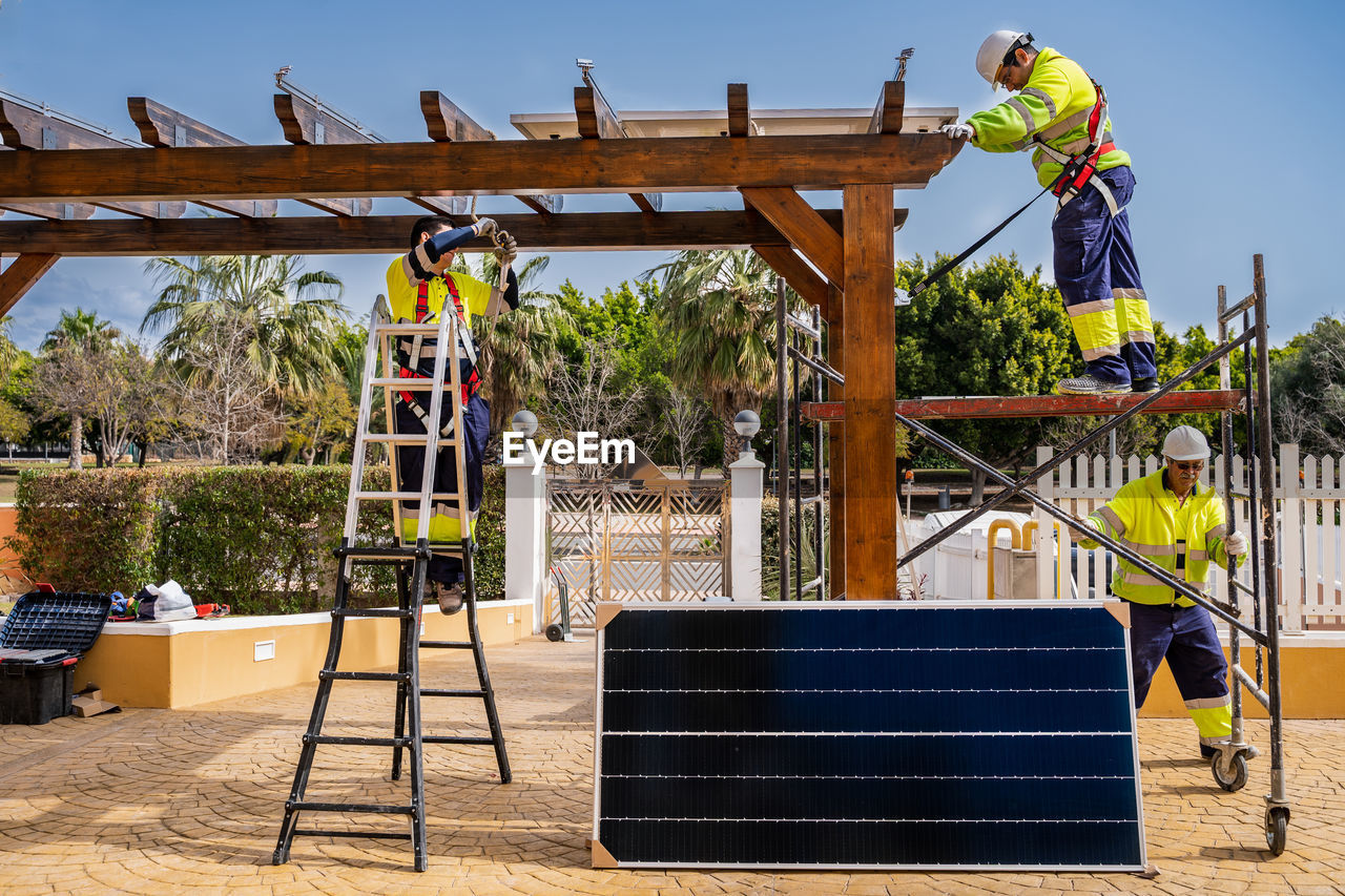 Group of workers in uniform and helmets installing photovoltaic panels on roof of wooden construction near house