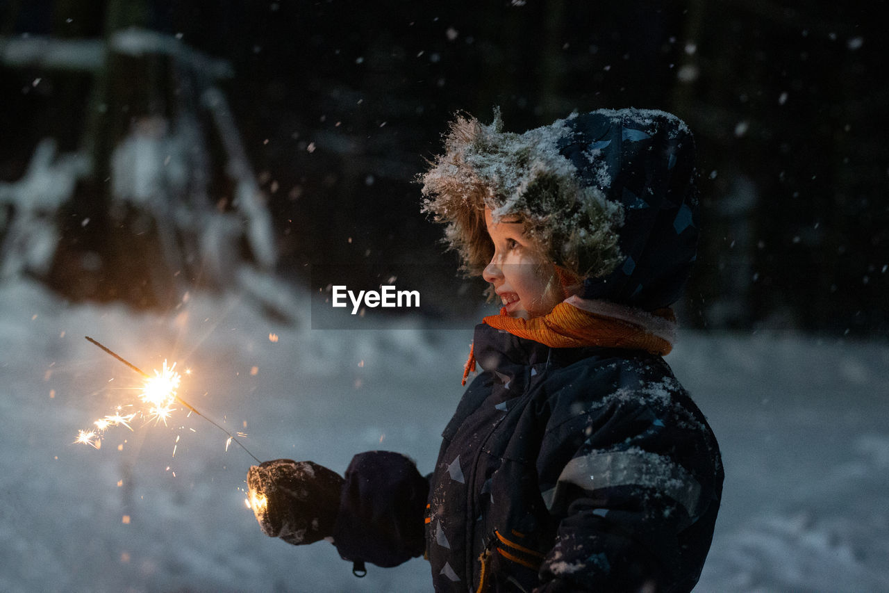 Toddler girl in winter clothes walking outside and holding sparkler in her hand. dark and snowy