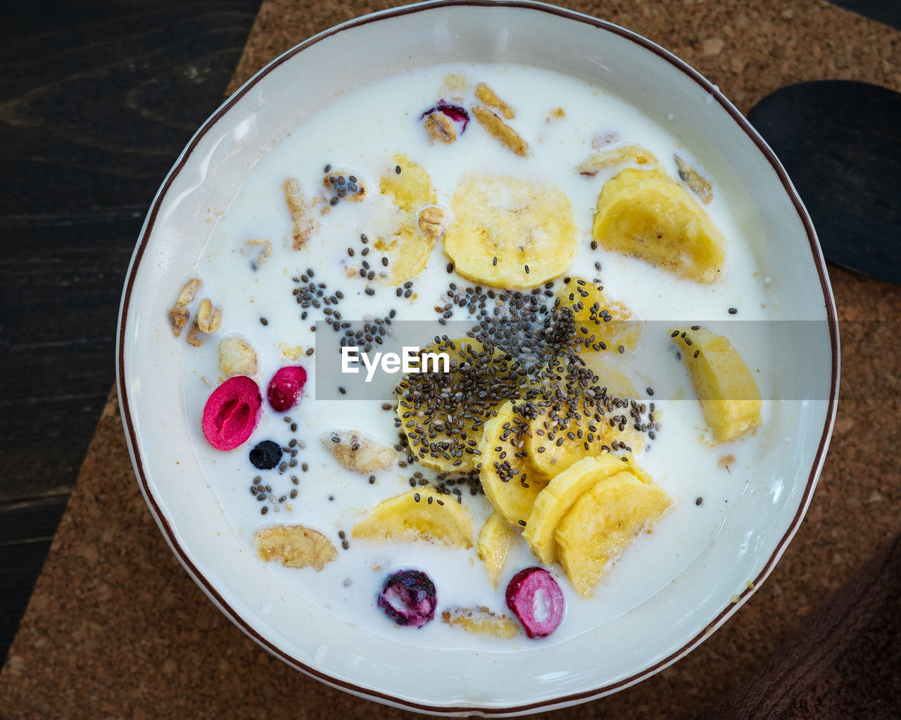 Fresh granola, banana with milk in a white bowl on brown background. sprinkled with chia seeds.