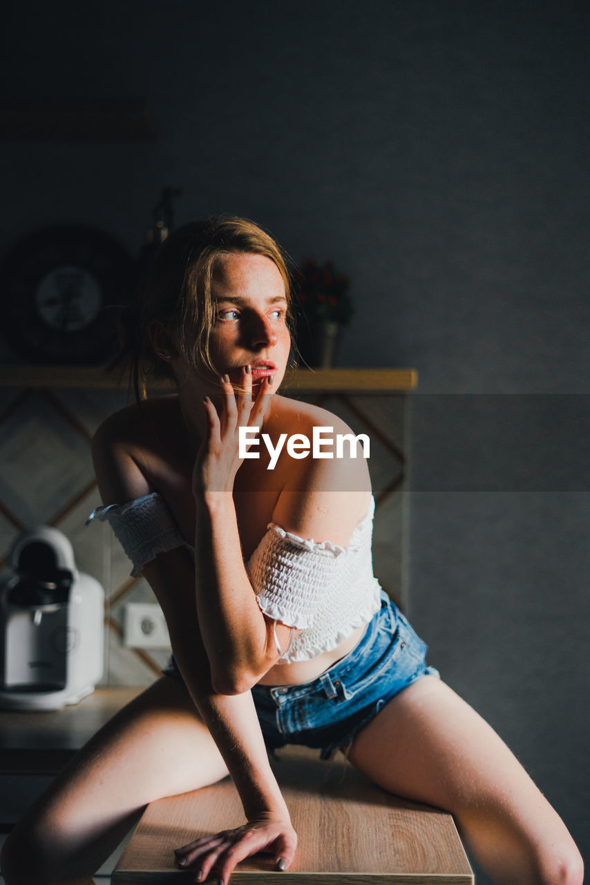 Unemotional young female in shorts and top with bare shoulders sitting on kitchen counter and looking away calmly